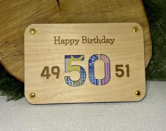 May include: A wooden card with the words "Happy Birthday" and the number "50" in the center. The number "50" is made of a cutout of a banknote. The numbers "49" and "51" are on either side of the number "50".