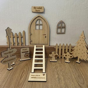 以下が含まれることがあります： 「Hier wohnt Schne」と書かれた木製の妖精のドア。ドアは開いており、梯子がドアまで続いています。また、木製のフェンス、郵便受け、街灯、