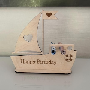 Könnte beinhalten: Ein Geschenkhalter in Form eines Holzsegelbootes mit der Aufschrift "Happy Birthday" auf dem Rumpf. Das Segel hat einen herzförmigen Ausschnitt und ein Herz auf der Flagge. Zwei aufgerollte Notizen sind sichtbar.