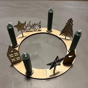 May include: A wooden Advent wreath with four candle holders, two stars, a Christmas tree, and two houses. The wreath is decorated with the word "Advent" written twice.