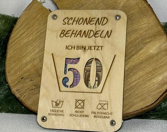 May include: A wooden sign with the text "SCHONEND BEHANDELN ICH BIN JETZT 50" and laundry care symbols. The sign is decorated with a 50 euro bill.