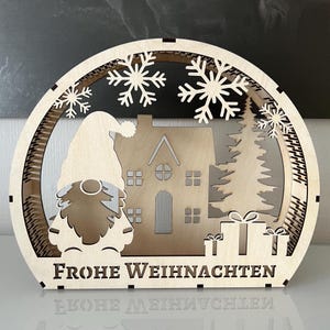 Può includere: Decorazione natalizia in legno tagliata al laser. Il design include uno gnomo, una casa, fiocchi di neve e la frase tedesca "Frohe Weihnachten". Sono inclusi anche dei regali.