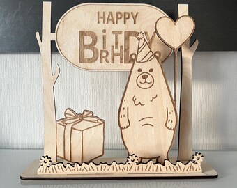 Peut inclure: Carte d'anniversaire en bois avec un ours portant un chapeau de fête et tenant un ballon en forme de cœur. La carte dit "Joyeux anniversaire" et a une boîte cadeau et des arbres en arrière-plan.