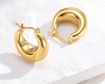 May include: Two gold-colored, chunky hoop earrings. The earrings have a smooth, polished surface and a rounded, hollow design. The earrings are displayed on a white surface, with soft lighting creating highlights and shadows.