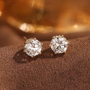 Puede incluir: Un par de pendientes de oro con grandes piedras preciosas redondas y transparentes. Los pendientes están engastados en un engaste de bisel dorado y se exhiben sobre una superficie marrón. Las piedras preciosas brillan, reflejando la luz.