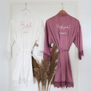 Könnte beinhalten: Zwei Satin-Roben mit Spitzenbesatz, eine weiße mit "Bride Maria" in Pink gestickt, die andere staubig-rosa mit "Bridesmaid Helena" in Weiß gestickt.