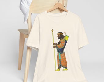 Può includere: Una t-shirt color crema con una grafica di un uomo in una tunica colorata che tiene una lancia. L'uomo ha una barba nera e la pelle marrone. La grafica è in stile cartone animato.