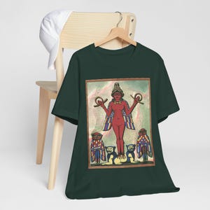 以下が含まれることがあります： 濃い緑色のTシャツに、腕を広げ、王冠をかぶった女性のカラフルなイラストが描かれています。その女性は、翼のある小さな人物3人に囲まれています。イラストは、白い縁取りのある薄い青色の背景に描かれています。