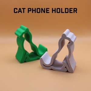 Op de afbeelding: Twee kattenvormige telefoonhouders, een groene en een witte, worden getoond op een lichtbruine ondergrond. De tekst "CAT PHONE HOLDER" wordt boven de houders weergegeven.
