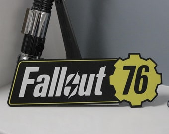 May include: A black and yellow Fallout 76 sign with white text and a yellow gear logo. A Vault-Tec figurine in a blue and yellow jumpsuit stands nearby. A black and silver lightsaber is in the background.