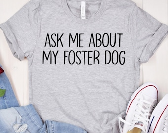 Puede incluir: Una camiseta gris con el texto "Ask me about my foster dog" impreso en ella. La camiseta está colocada sobre una superficie de madera blanca con un par de pantalones vaqueros azules, una rosa roja y zapatillas blancas.
