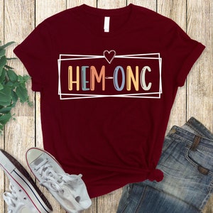 Puede incluir: Una camiseta color granate con un contorno blanco de un coraz&oacute;n y una caja con el texto "HEM-ONC" en letras de colores.