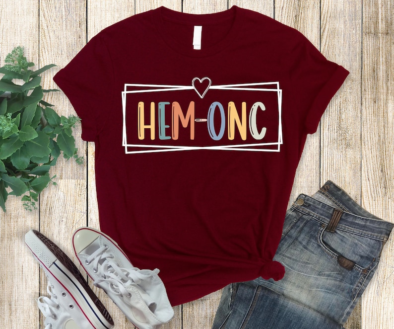 Puede incluir: Una camiseta color granate con un contorno blanco de un coraz&oacute;n y una caja con el texto "HEM-ONC" en letras de colores.