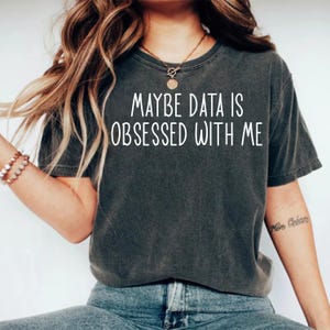 May include: Dark gray t-shirt with the text "MAYBE DATA IS OBSESSED WITH ME" in white. The shirt is a short-sleeved, casual style. The model is wearing blue jeans and a necklace.