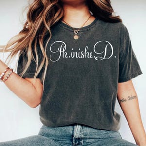 May include: Dark gray t-shirt with the cursive text "Th.inishe.D." in white. The shirt has a vintage, worn look. The model is wearing blue jeans and a necklace.