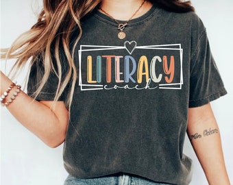 May include: A dark grey t-shirt with a white outline box around the text "Literacy Coach" in rainbow colors with a small heart above the text.