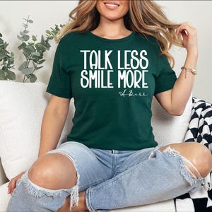 Pu&ograve; includere: Una maglietta verde scuro con la scritta bianca "TALK LESS SMILE MORE". La maglietta presenta anche la firma "A. Burr" in corsivo. La persona indossa jeans blu strappati.