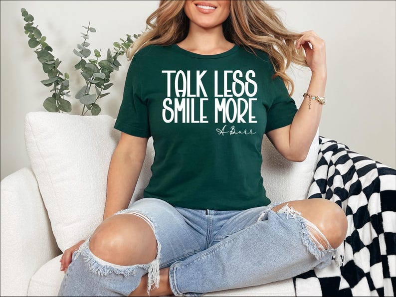 Pu&ograve; includere: Una maglietta verde scuro con la scritta bianca "TALK LESS SMILE MORE". La maglietta presenta anche la firma "A. Burr" in corsivo. La persona indossa jeans blu strappati.