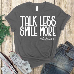 Pu&ograve; includere: T-shirt grigio scuro con la scritta bianca "TALK LESS SMILE MORE" e la firma corsiva "A Burr". La maglietta &egrave; abbinata a jeans blu e scarpe da ginnastica bianche.
