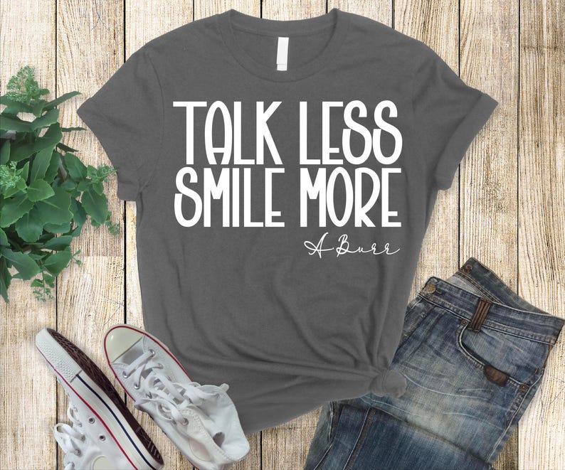 Pu&ograve; includere: T-shirt grigio scuro con la scritta bianca "TALK LESS SMILE MORE" e la firma corsiva "A Burr". La maglietta &egrave; abbinata a jeans blu e scarpe da ginnastica bianche.