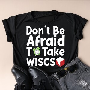 May include: Black t-shirt with white text that reads "Don't Be Afraid To Take WISC's". A green alarm clock and a red cube are next to the text. The shirt is laying on a pair of black shoes and a pair of denim shorts.