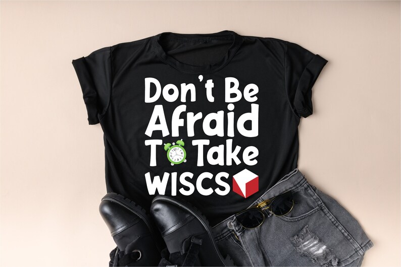 May include: Black t-shirt with white text that reads "Don't Be Afraid To Take WISC's". A green alarm clock and a red cube are next to the text. The shirt is laying on a pair of black shoes and a pair of denim shorts.