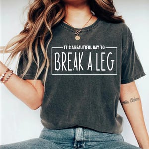 May include: Dark grey t-shirt with the text "IT'S A BEAUTIFUL DAY TO BREAK A LEG" in a white rectangular box. The shirt is a crew neck style and appears to be made of a soft, comfortable material. The shirt is worn by a person.