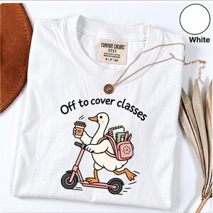 May include: A white t-shirt featuring a graphic of a white goose riding a pink scooter. The goose wears a pink backpack filled with notebooks and pens while holding a coffee cup. The text "Off to cover classes" is printed above the goose.