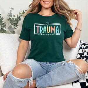 May include: A dark green t-shirt with a white frame and the text "Trauma Therapist" in rainbow colors. A small pink heart is above the text.
