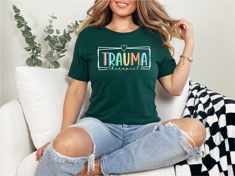 May include: A dark green t-shirt with a white frame and the text "Trauma Therapist" in rainbow colors. A small pink heart is above the text.