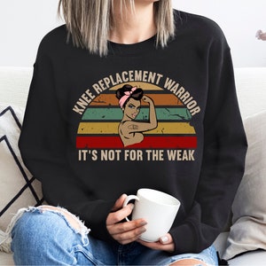May include: A black sweatshirt with a vintage-style graphic of a woman with a pink headband and the text "Knee Replacement Warrior It's Not For The Weak".