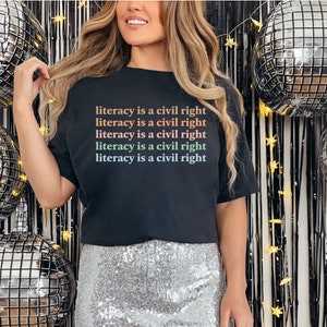 May include: A black t-shirt with the text "literacy is a civil right" repeated five times in rainbow colours. The shirt is worn by a person standing in front of a silver and black disco ball backdrop.