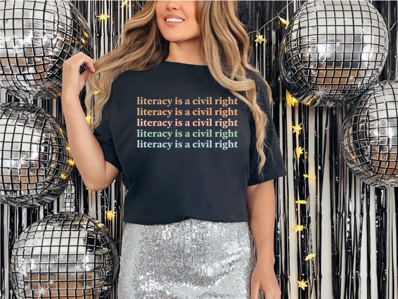May include: A black t-shirt with the text "literacy is a civil right" repeated five times in rainbow colours. The shirt is worn by a person standing in front of a silver and black disco ball backdrop.
