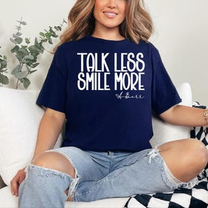 Pu&ograve; includere: T-shirt blu navy con la scritta bianca "TALK LESS SMILE MORE". La maglietta presenta anche una firma bianca. La modella indossa jeans blu strappati.