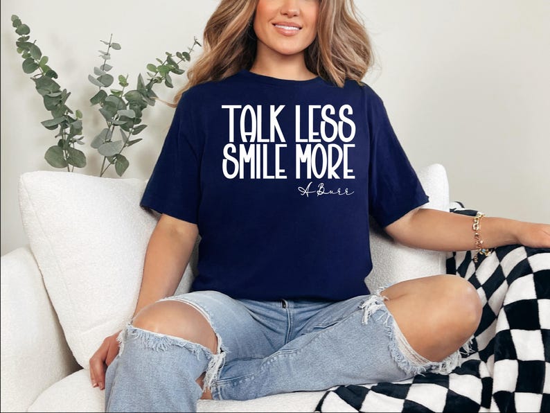 Pu&ograve; includere: T-shirt blu navy con la scritta bianca "TALK LESS SMILE MORE". La maglietta presenta anche una firma bianca. La modella indossa jeans blu strappati.