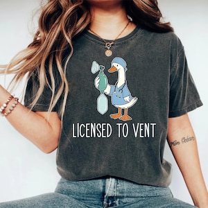 May include: Dark gray t-shirt featuring a cartoon goose in medical scrubs holding a ventilator. The goose wears a blue scrub cap and a stethoscope. The text "LICENSED TO VENT" is printed in white below the graphic. A casual, comfortable tee.