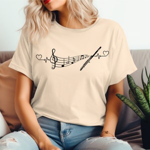 Może przedstawiać: Beżowa koszulka z czarnym nadrukiem fletu i nut muzycznych z linią bicia serca i sercami po obu stronach. Na koszulce widnieje napis "Music is my heartbeat".