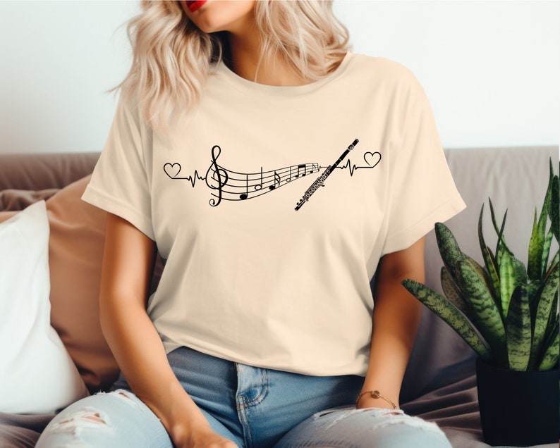 Może przedstawiać: Beżowa koszulka z czarnym nadrukiem fletu i nut muzycznych z linią bicia serca i sercami po obu stronach. Na koszulce widnieje napis "Music is my heartbeat".