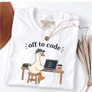 May include: A white t-shirt featuring a graphic of a goose wearing a grey cap and round glasses, sitting on a stool at a desk with a laptop, books, and a coffee cup. The text "off to code" is printed above the goose in a black sans-serif font.