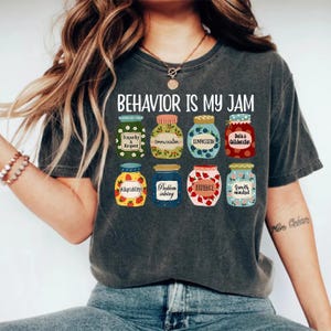 May include: Dark gray t-shirt with the text "BEHAVIOR IS MY JAM" above illustrations of jam jars. Each jar has a label with a different positive behavior, such as "Empathy & Respect" and "Patience".