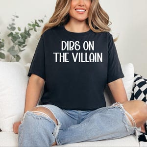 May include: Black t-shirt with the text "DIBS ON THE VILLAIN" in white block letters. The shirt is worn with light blue ripped jeans. The person is sitting on a white couch with a black and white checkered blanket.