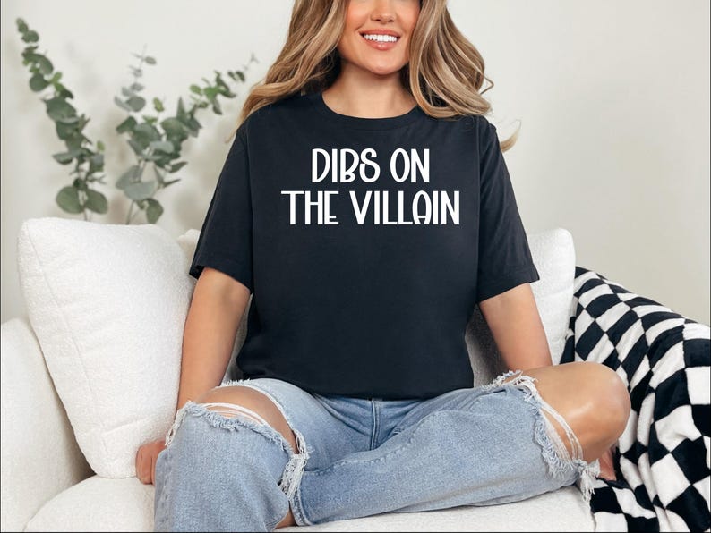 May include: Black t-shirt with the text "DIBS ON THE VILLAIN" in white block letters. The shirt is worn with light blue ripped jeans. The person is sitting on a white couch with a black and white checkered blanket.