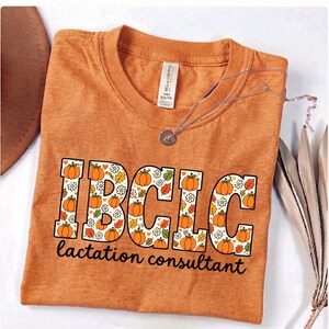 May include: A heathered orange t-shirt featuring the text IBCLC in large block letters filled with a pattern of orange pumpkins, white daisies, and autumn leaves. Below the letters, the text lactation consultant is printed in black script.
