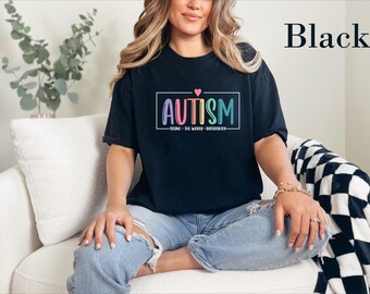 May include: A black short-sleeve t-shirt featuring a rectangular graphic with the word AUTISM in colorful letters, a small pink heart above, and the text SEEING THE WORLD DIFFERENTLY below, all enclosed in a white rectangular border.