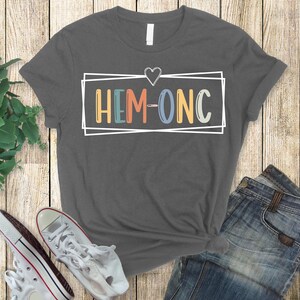 Puede incluir: Una camiseta gris oscuro con un coraz&oacute;n blanco y un rect&aacute;ngulo blanco con el texto "HEM-ONC" en letras de colores.