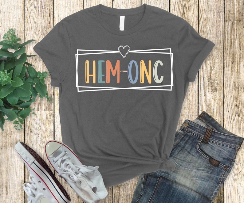 Puede incluir: Una camiseta gris oscuro con un coraz&oacute;n blanco y un rect&aacute;ngulo blanco con el texto "HEM-ONC" en letras de colores.
