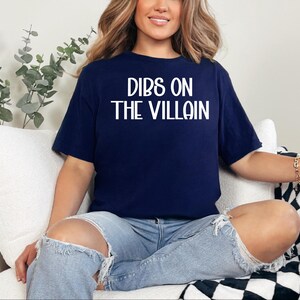 May include: Navy blue t-shirt with the white text "DIBS ON THE VILLAIN". The shirt is a short-sleeved crew neck. The person is wearing light blue ripped jeans. The background includes a white couch and a black and white checkered blanket.