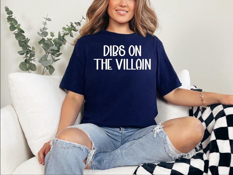 May include: Navy blue t-shirt with the white text "DIBS ON THE VILLAIN". The shirt is a short-sleeved crew neck. The person is wearing light blue ripped jeans. The background includes a white couch and a black and white checkered blanket.
