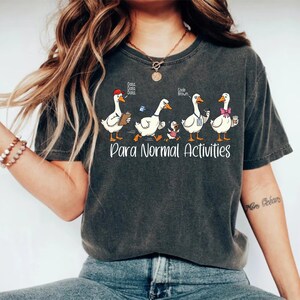 May include: A charcoal gray short-sleeve t-shirt featuring a graphic of five white geese. The text reads "Data, Data, Data," "Code Brown," and "Para Normal Activities." The geese are depicted wearing various accessories including a red cap and a pink bow.