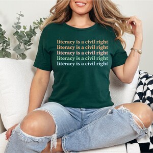May include: A woman wearing a dark green t-shirt with the text "literacy is a civil right" repeated five times in a gradient of orange, pink, and blue. She is sitting on a white sofa with a black and white checked throw blanket.